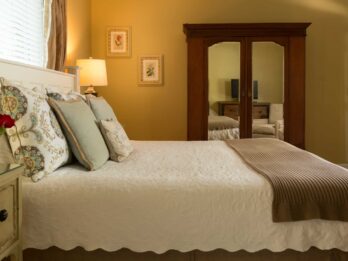 A room at the Chanticleer Inn shows a large bed with white linens, modern throw pillows, an antique side table with fresh roses, and a mirrored dresser.