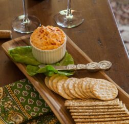 Pimento cheese and crackers rest on a wooden tray with two wine glasses in the background.